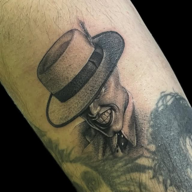 Black-and-grey tattoo of a sinister smiling figure in a wide-brimmed hat